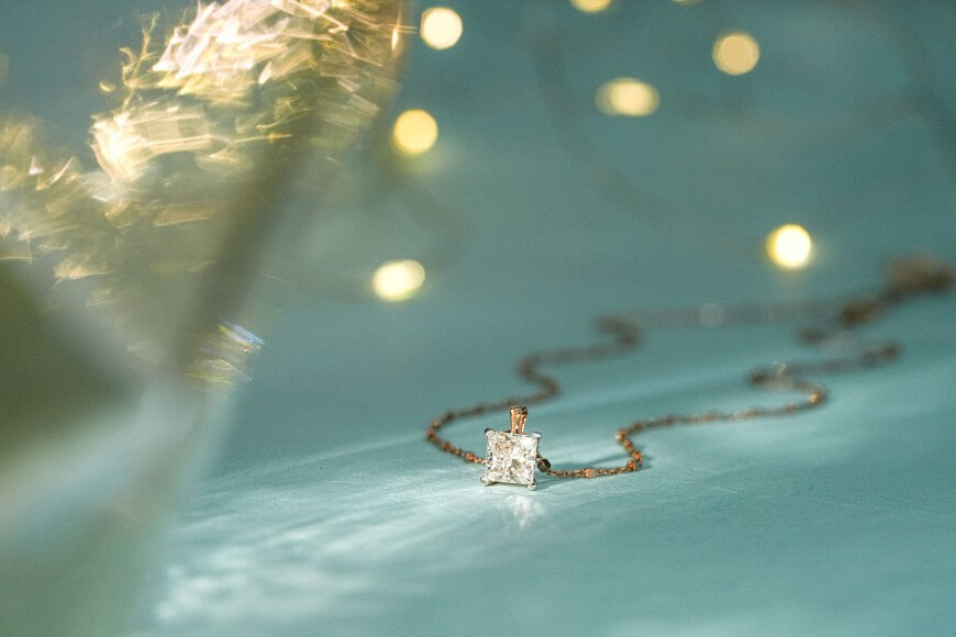 A gold necklace with a square-cut diamond pendant lies on a light blue surface, with blurred golden lights and bokeh effects in the background, creating a dreamy, elegant atmosphere.