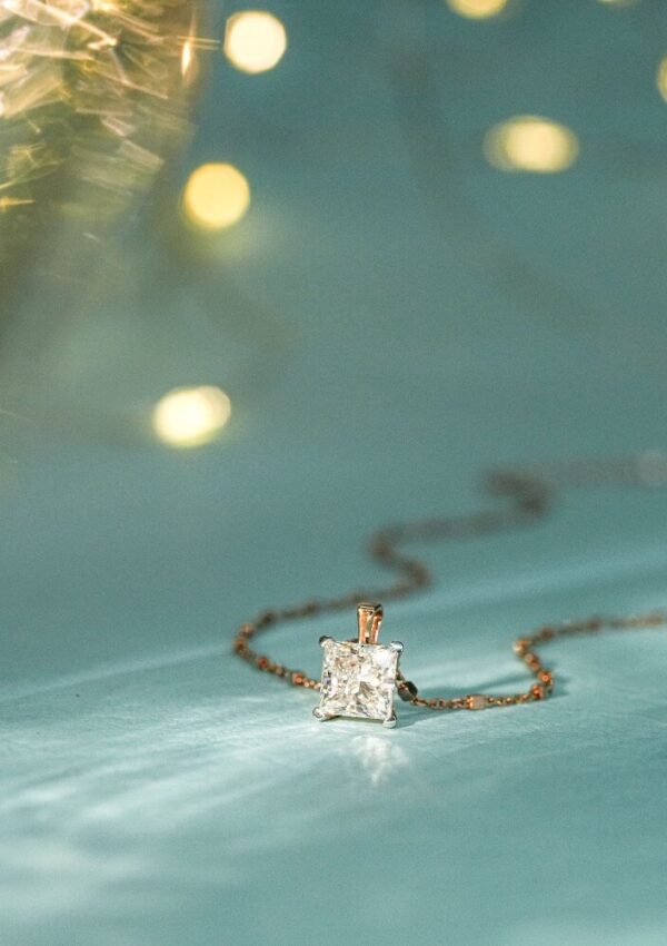 A delicate gold chain necklace with a square-cut diamond pendant is displayed on a light blue surface, with soft yellow lights blurred in the background.