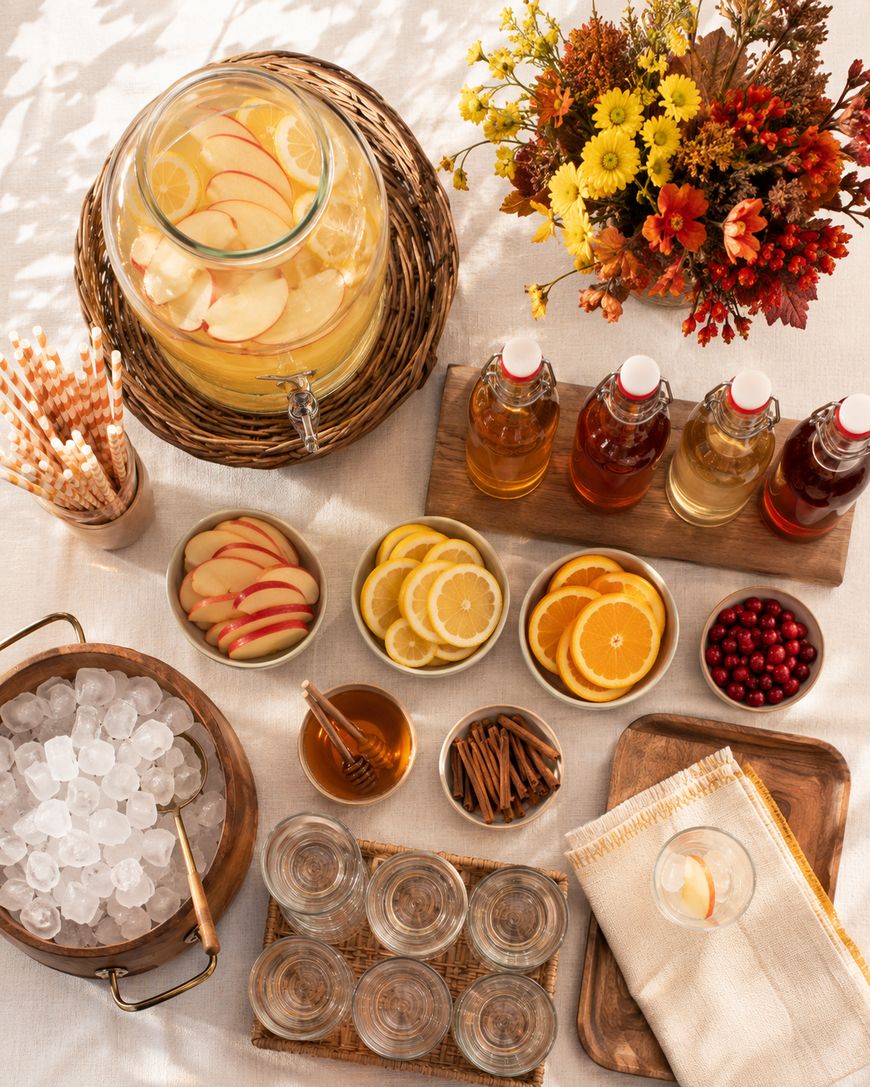 Overhead view of a fall drink station with apple cider, fruit slices, syrups, ice, glasses, cinnamon sticks, cranberries, honey, and striped straws on a light tablecloth, with a bouquet of autumn flowers.