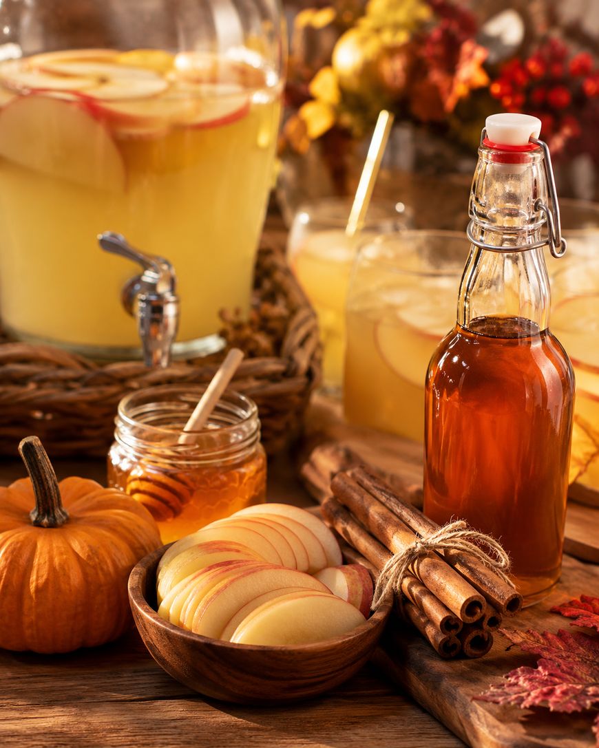 A fall-themed setup with apple cider in a glass dispenser, a bottle, sliced apples, cinnamon sticks, honey, a small pumpkin, and autumn leaves, all arranged on a wooden table.