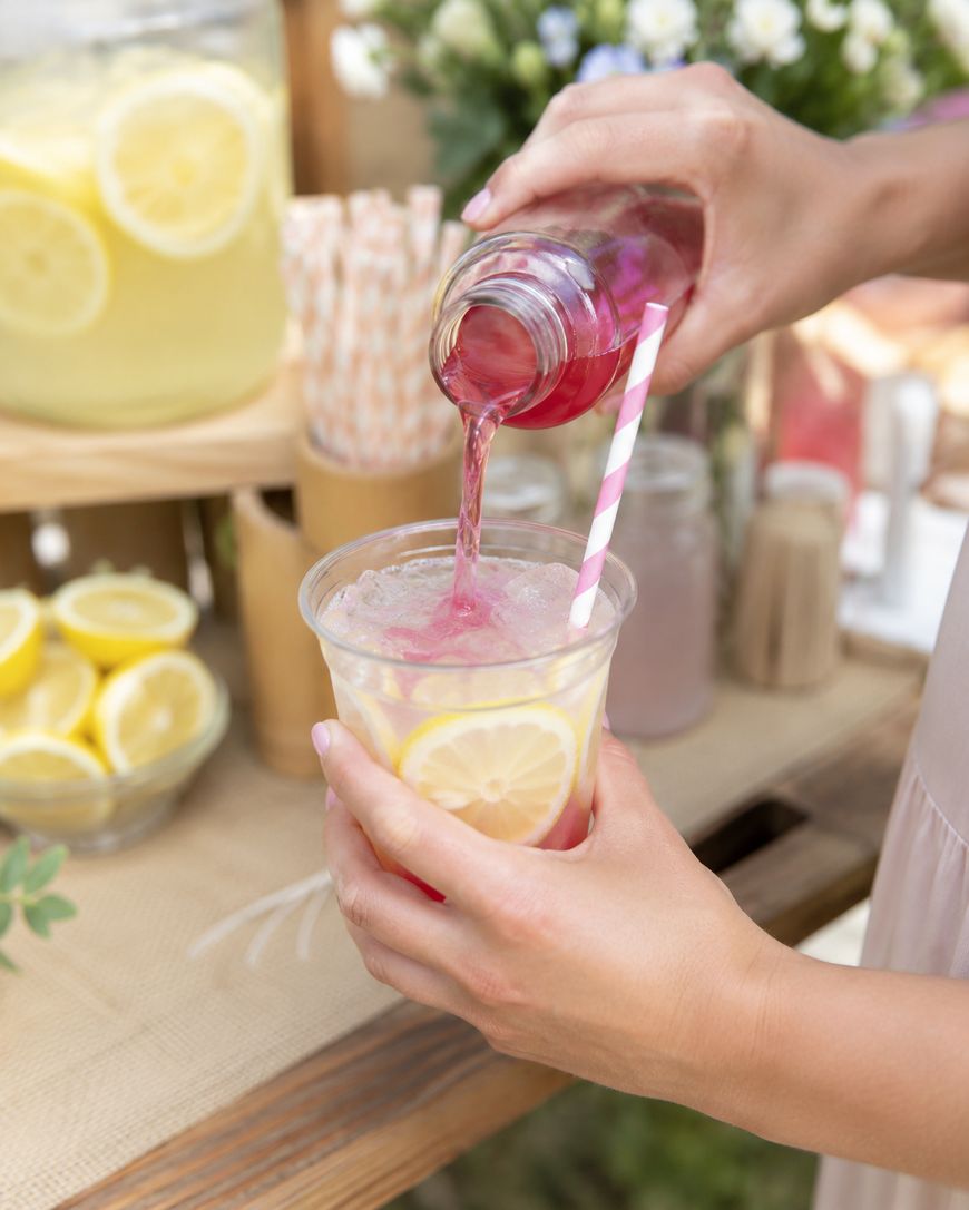 A person pours pink lemonade into a clear cup with lemon slices and a striped straw. A large pitcher of lemonade, lemon slices, and straws are visible in the background, suggesting a refreshing outdoor setting.
