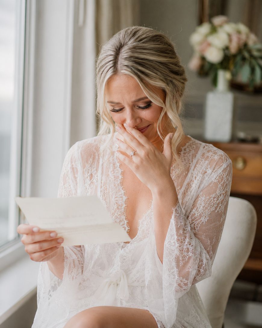 A bride in a lace robe sits by a window, smiling and wiping tears while reading a letter. A bouquet of flowers is blurred in the background.
