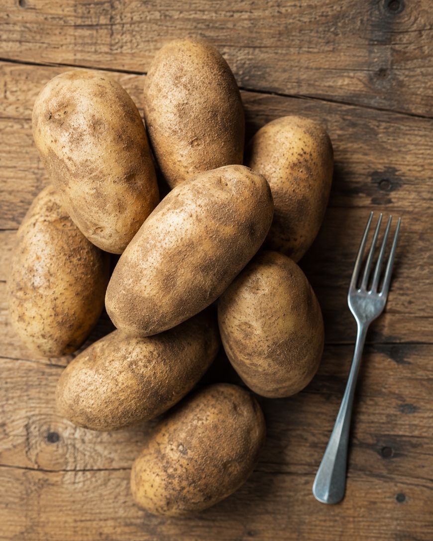 Eight unwashed russet potatoes are arranged on a rustic wooden surface next to a metal fork.