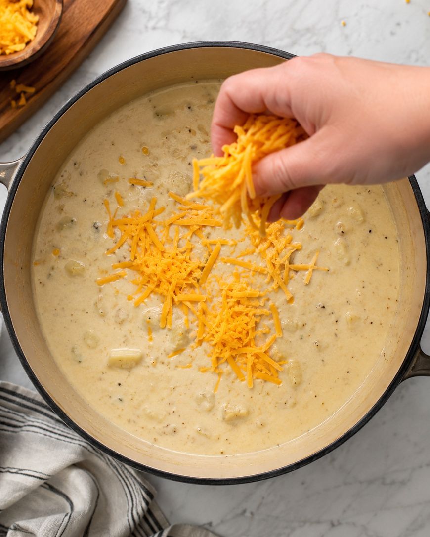 A hand sprinkles shredded cheddar cheese into a pot of creamy soup, likely potato soup, on a marble countertop with a striped cloth nearby.