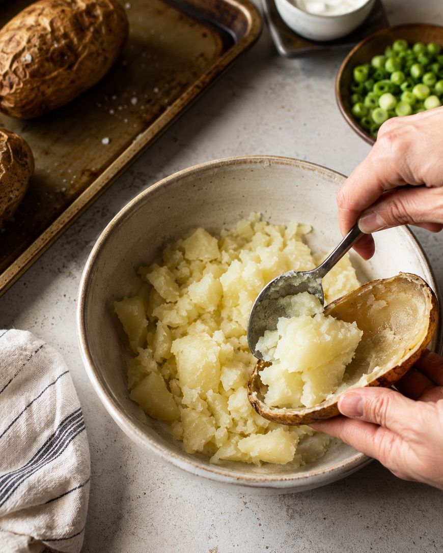 Hands scooping mashed baked potato from a bowl back into a potato skin, with baked potatoes, green onions, and a towel nearby on a countertop.