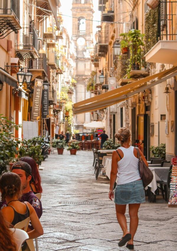 A woman in a white tank top and denim skirt walks down a narrow, sunny European street lined with cafes, plants, and colorful buildings. Several people are seated at tables, and a clock tower rises in the distance.