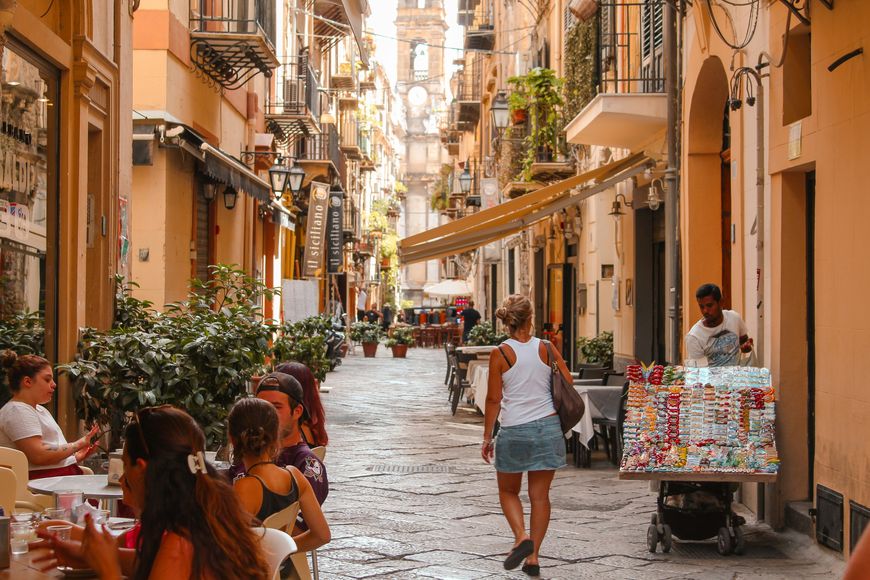 People sit at outdoor café tables along a narrow, cobblestone street lined with shops and plants. A woman walks past a man pushing a cart filled with colorful items. The atmosphere is lively and sunlit.