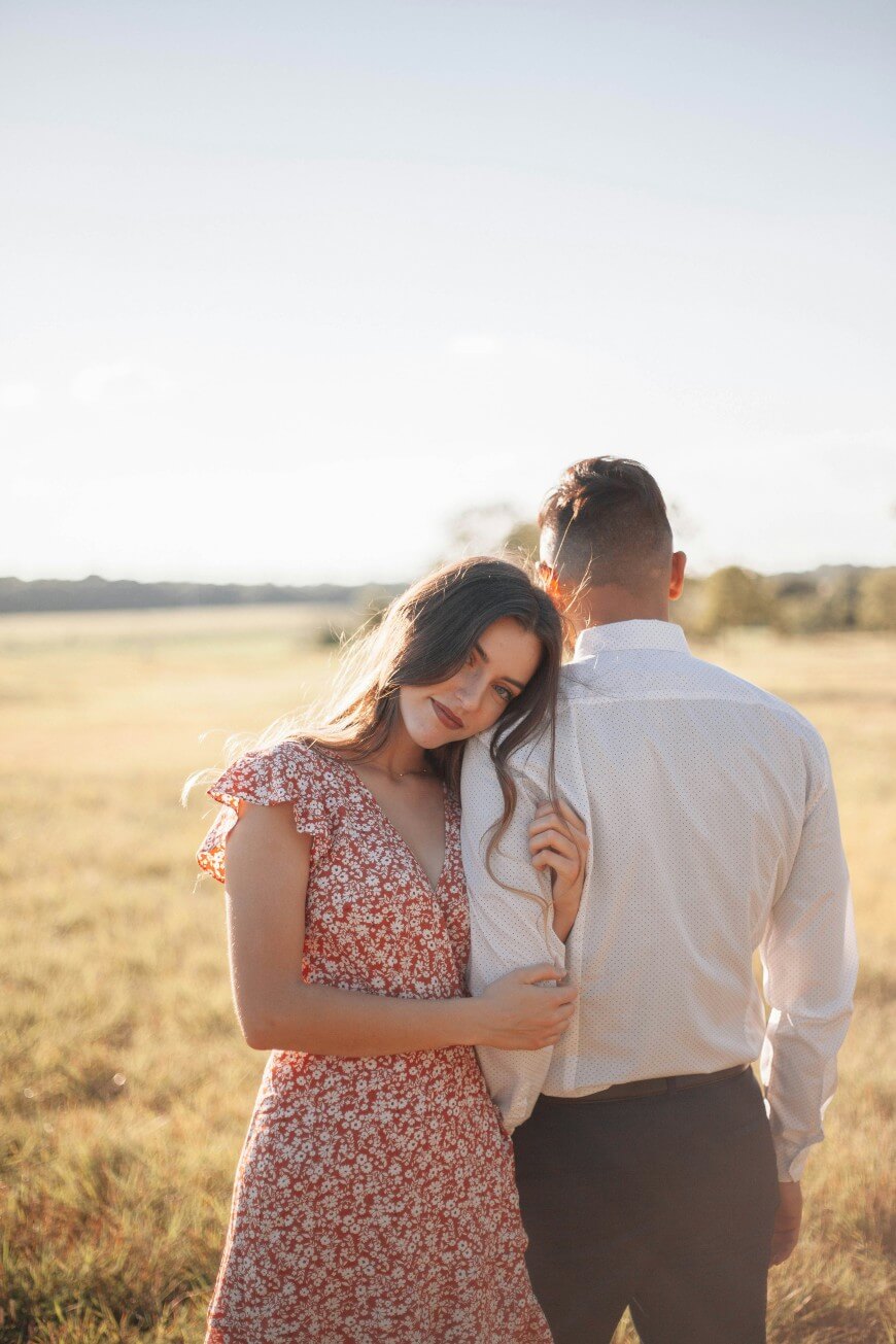 A woman in a floral dress leans her head on a mans shoulder while holding his arm, standing together in a sunlit field.