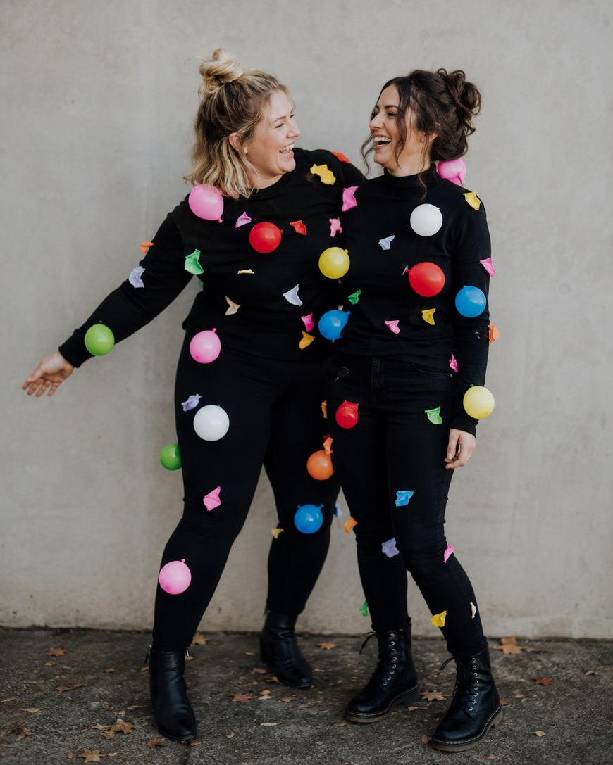 Two people in black outfits covered with colorful balloons and confetti pieces stand smiling and facing each other in front of a plain wall, with arms out. Both are wearing black boots and appear happy.