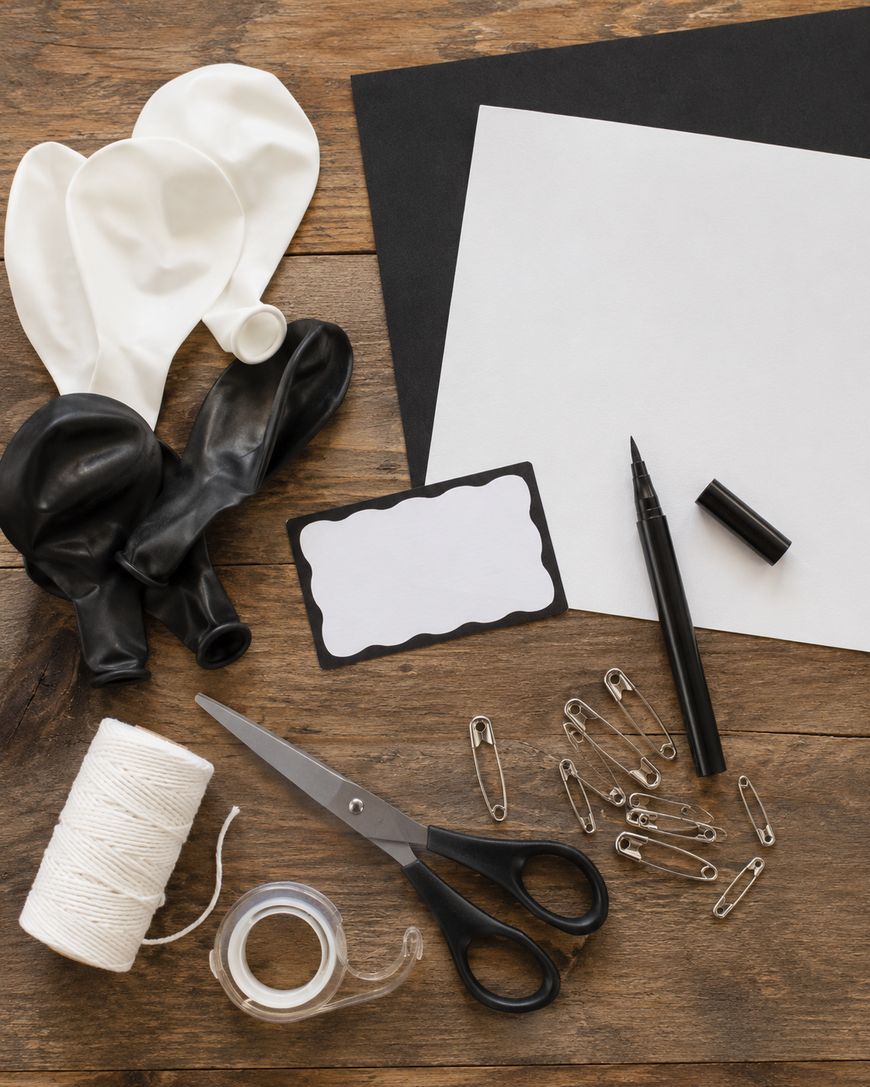 Black and white balloons, blank papers, a marker, scissors, string, safety pins, tape, and a label card are arranged on a wooden surface, suggesting a craft or party preparation scene.