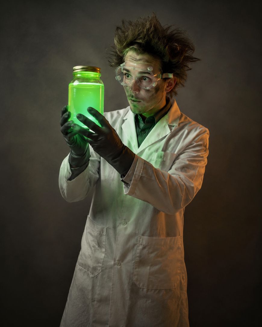 A scientist with messy hair and goggles, wearing a white lab coat and gloves, holds a glowing green jar and looks at it with a surprised expression.