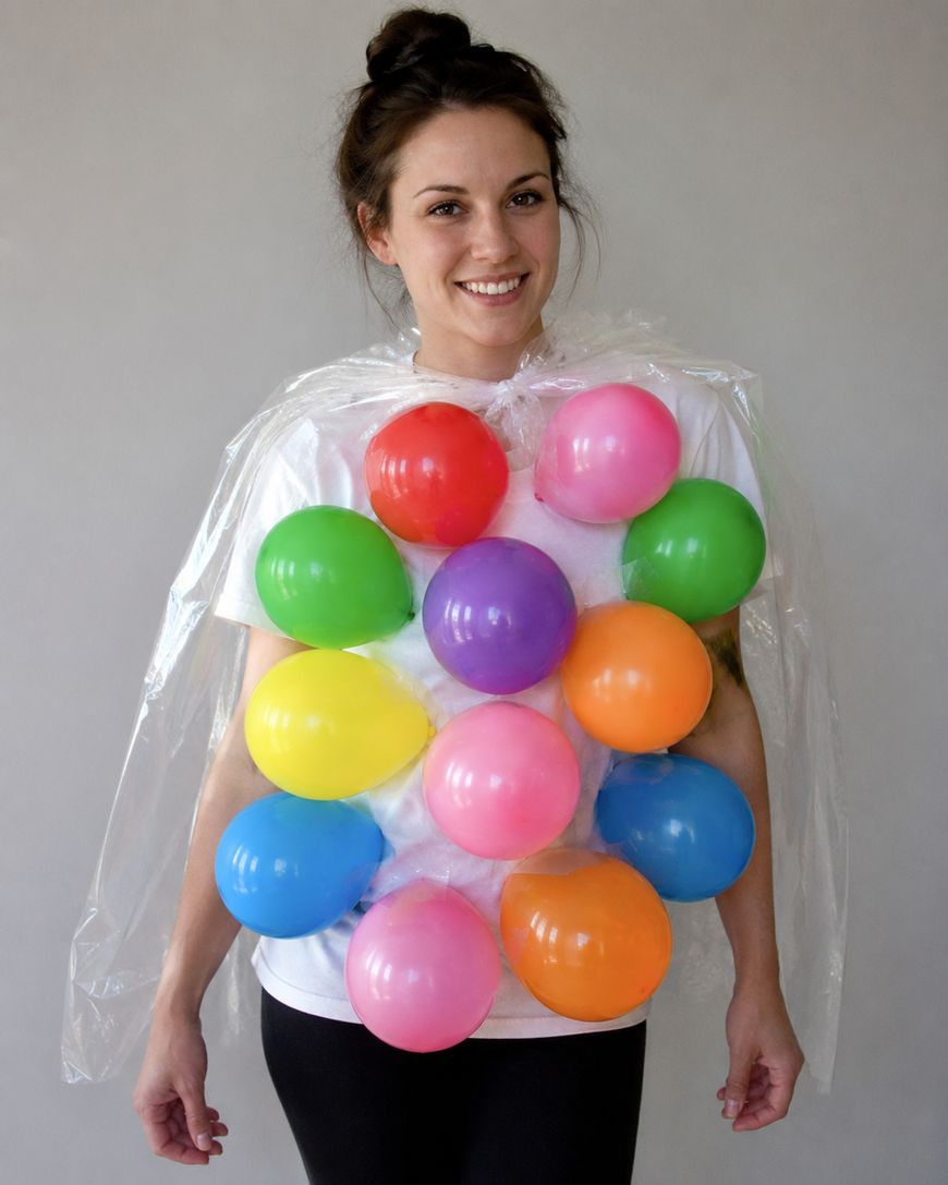 Smiling person wearing a homemade costume with colorful balloons attached to a clear plastic poncho, resembling a bunch of grapes or bubble wrap, standing against a plain background.