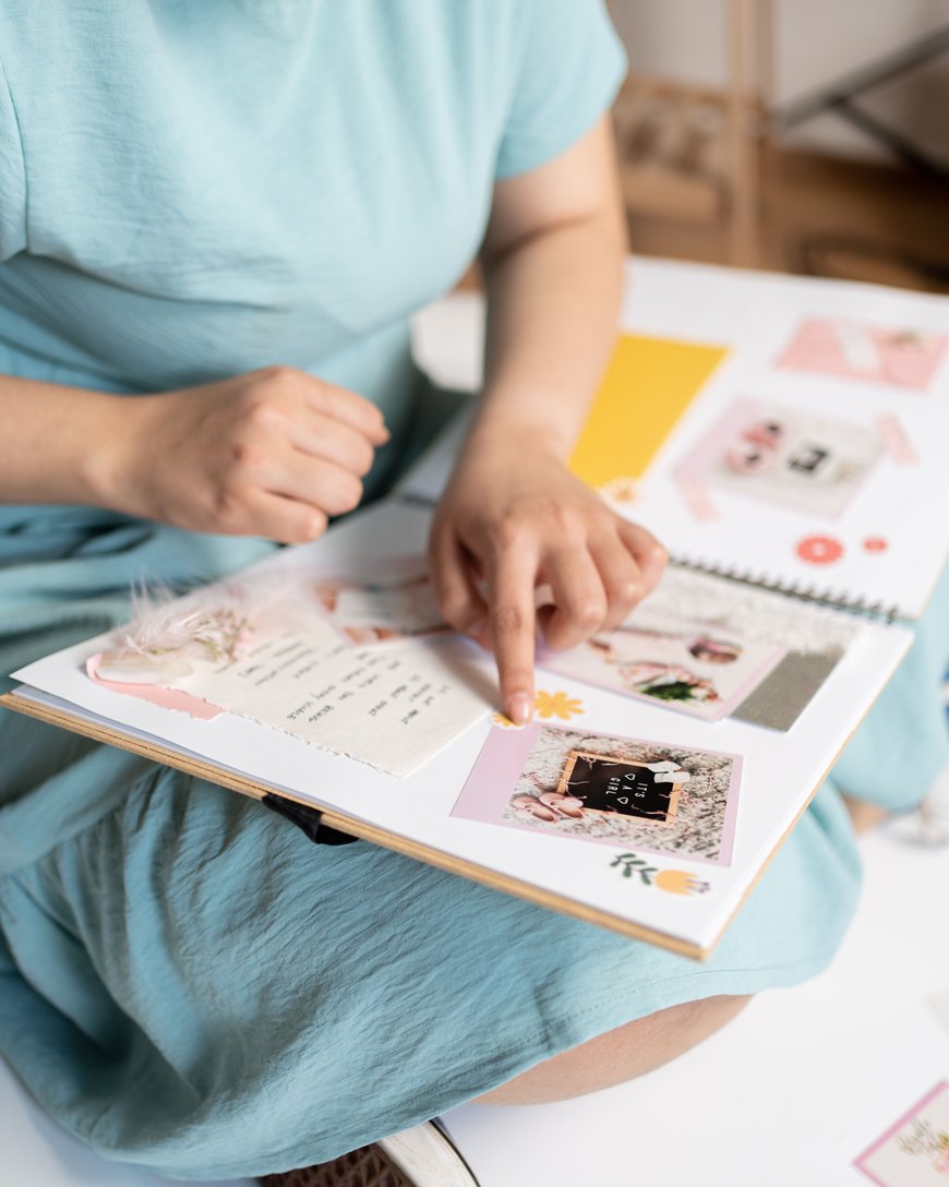 A person in a light blue dress sits and points to a photo in a scrapbook filled with pictures, notes, and decorative elements.