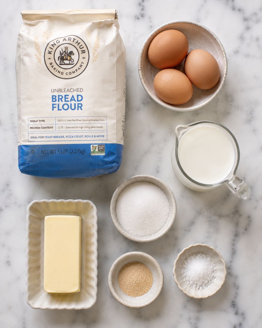 A bag of bread flour, three eggs in a bowl, a pitcher of milk, a dish of butter, and small bowls of sugar, salt, and yeast are arranged on a white marble surface.
