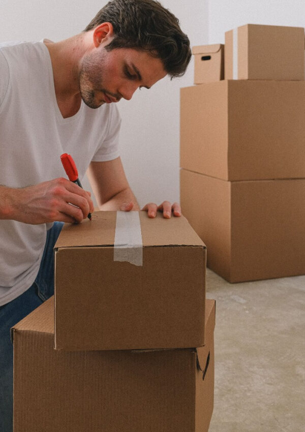 A man in a white t-shirt labels a cardboard box with a red marker in a room with stacked moving boxes on a light-colored floor.