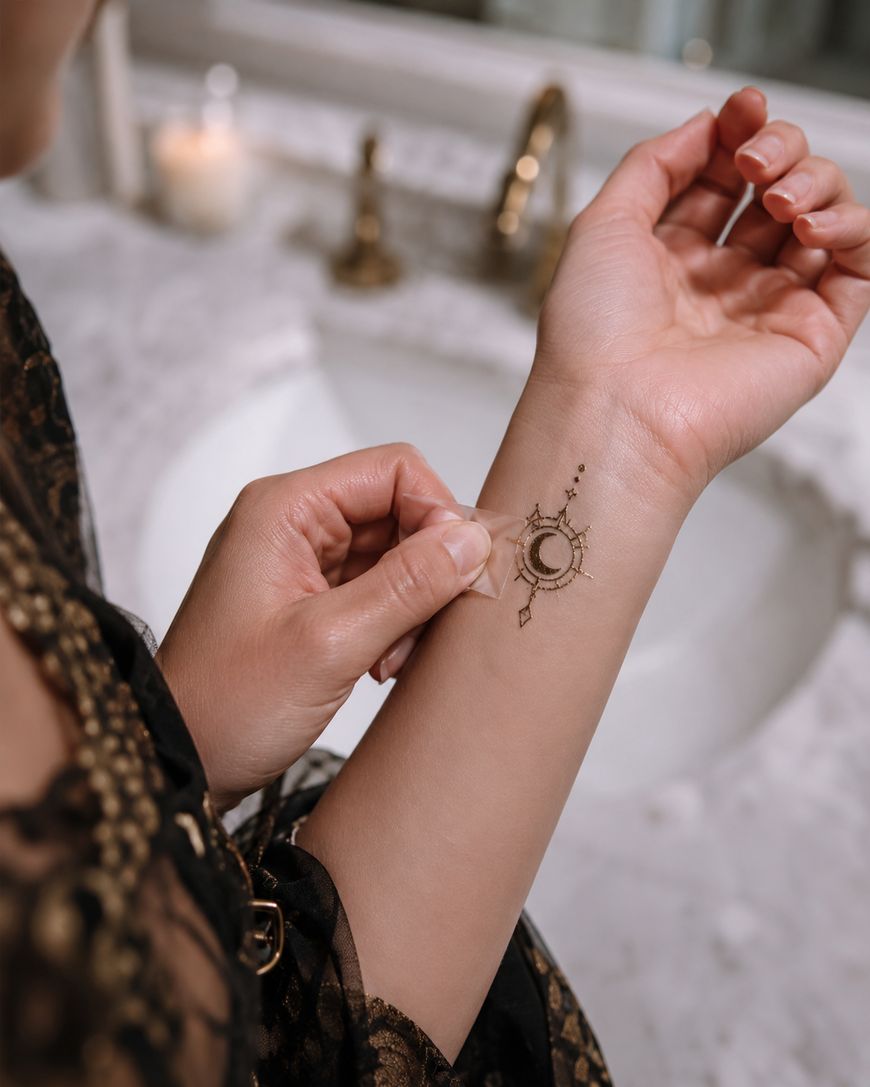 A person peels a temporary tattoo with a moon and geometric design from their forearm over a marble bathroom sink.