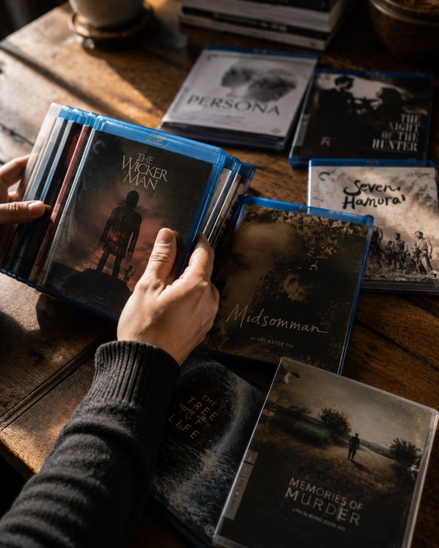 A person selects a Blu-ray of The Wicker Man from a wooden table covered with various Blu-ray and DVD cases, including Midsommar, Persona, and Memories of Murder.