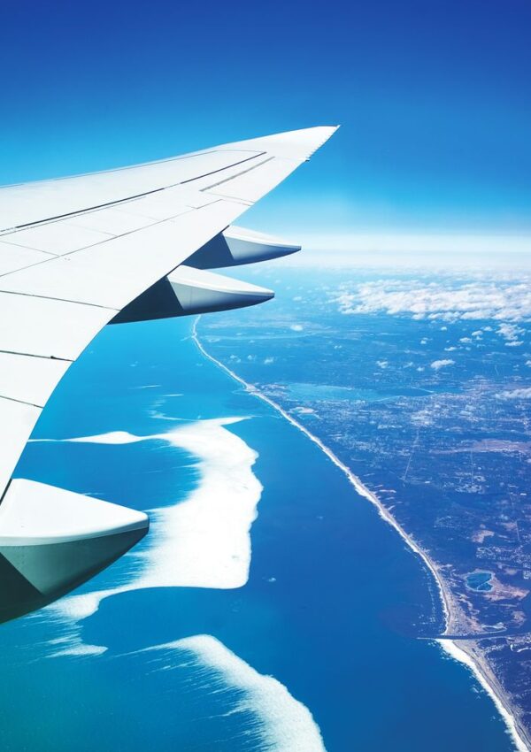 A view from an airplane window showing the wing above a coastline, with blue ocean, white waves, and land meeting the horizon under a clear sky.