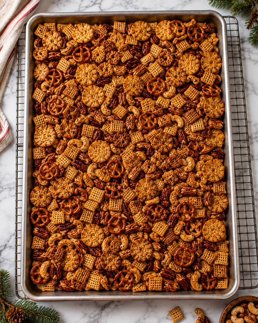 A large baking sheet filled with homemade snack mix, including pretzels, Chex cereal, nuts, and small crackers, sits on a cooling rack over a marble counter.