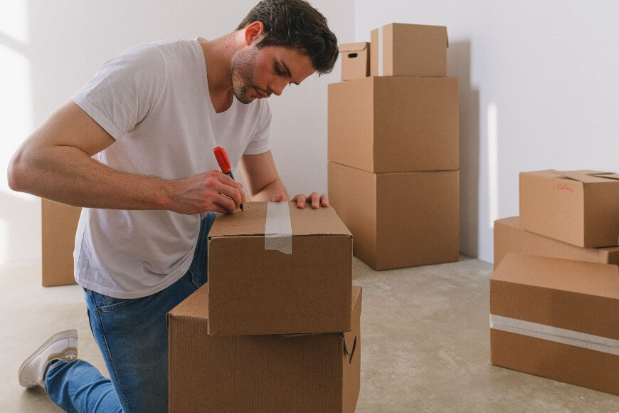 A man kneels on the floor and labels a cardboard moving box with a red marker. Several other cardboard boxes are stacked around him in a brightly lit, empty room.