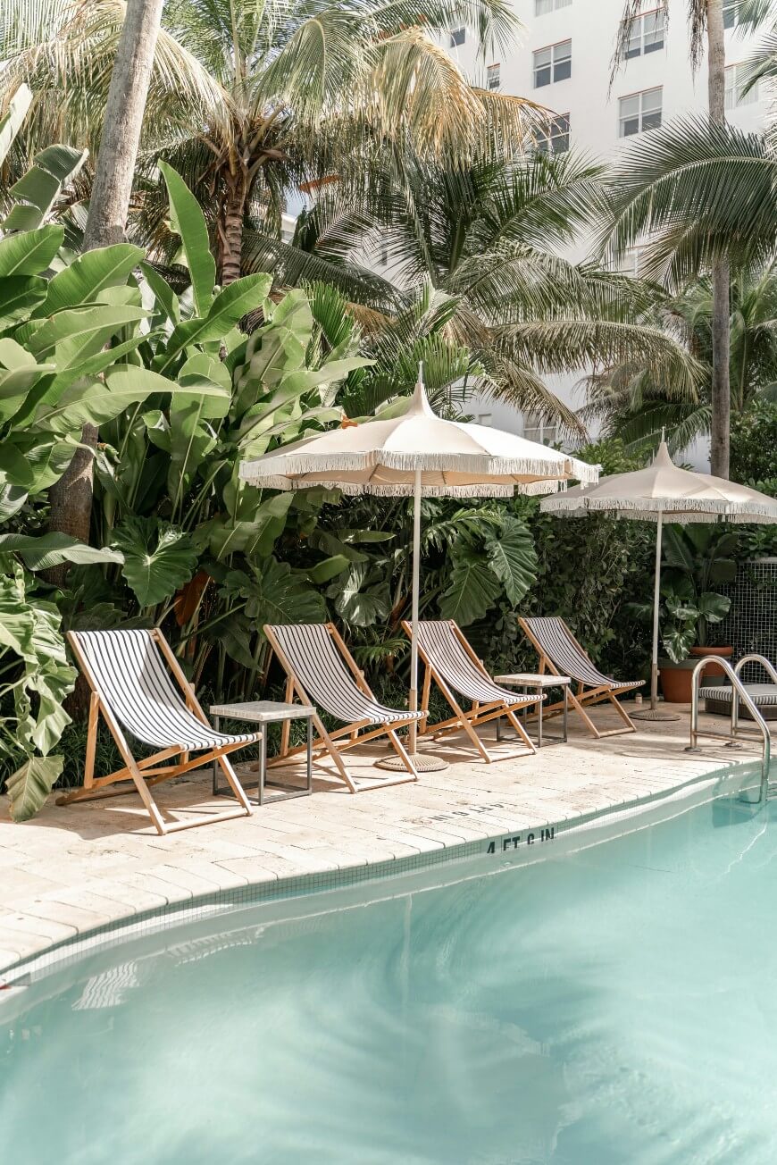 Poolside scene with striped lounge chairs and white umbrellas, surrounded by lush green tropical plants and palm trees. The water in the pool is calm, and a modern building is visible in the background.