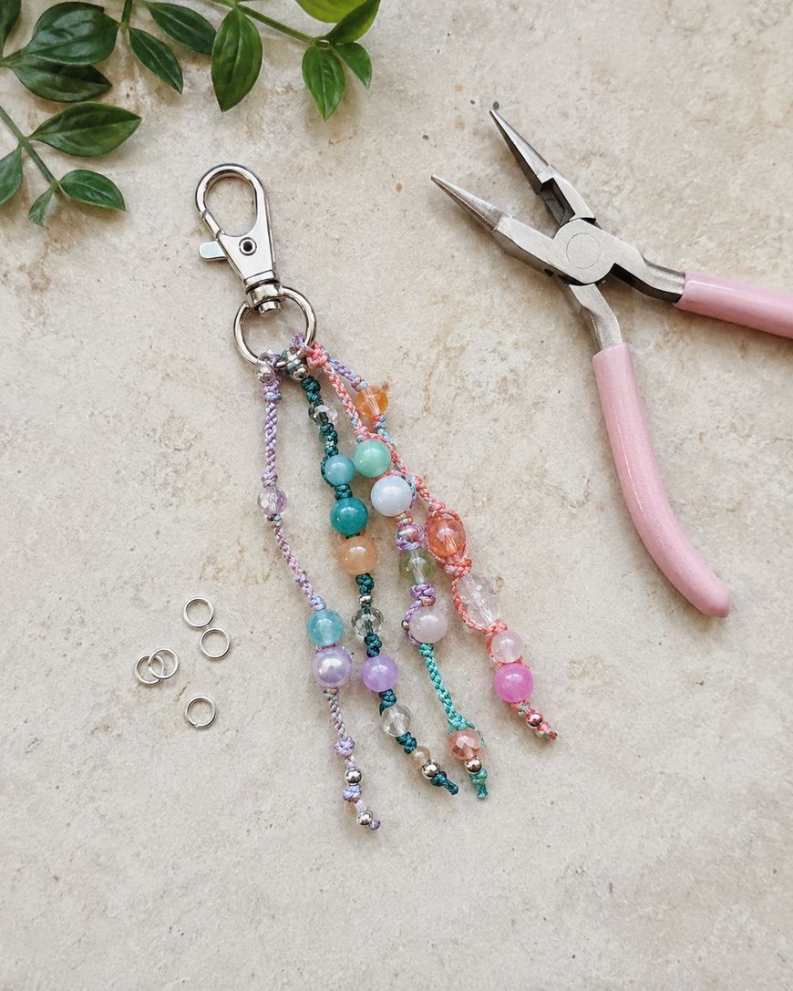 A beaded keychain with colorful beads, a pink-handled plier, four jump rings, and some green leaves on a beige surface.