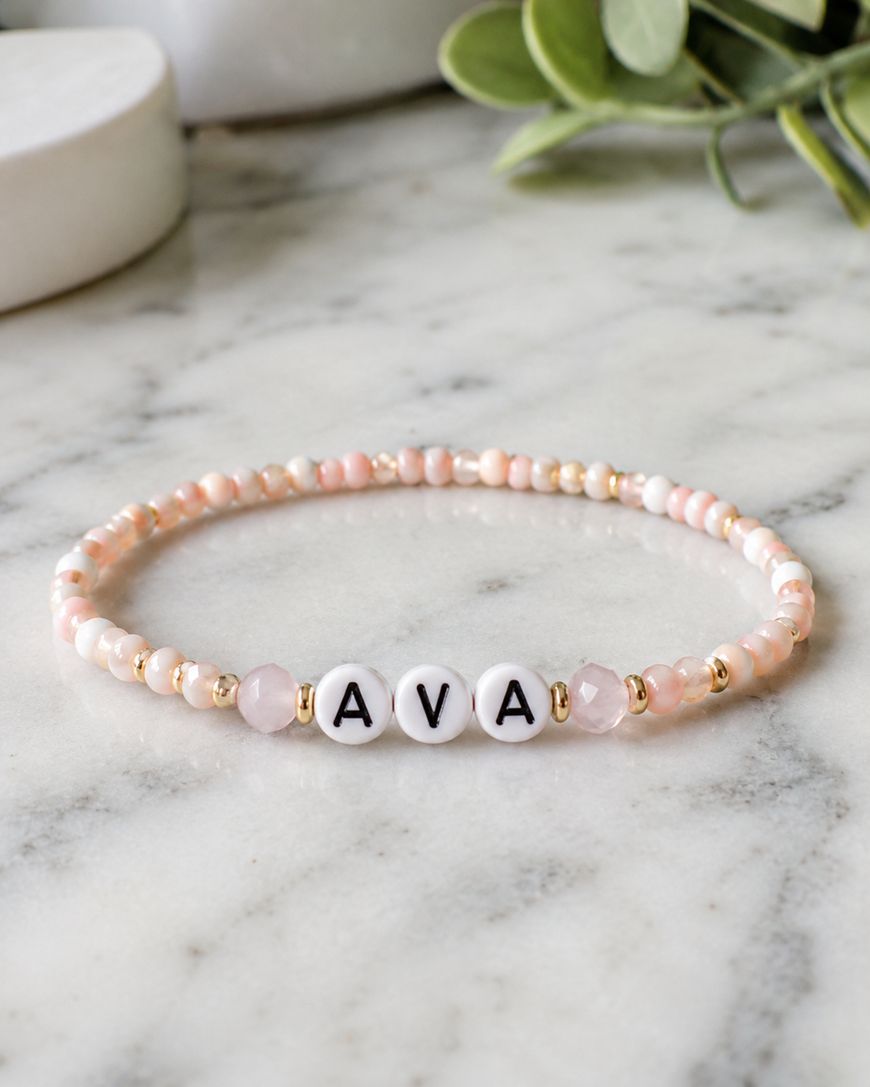 A beaded bracelet with pink and gold beads, featuring three white round beads in the center spelling AVA in black letters, displayed on a marble surface with green leaves in the background.