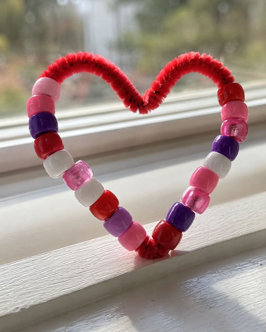 A heart shape made from a red pipe cleaner and colorful beads, including pink, red, white, and purple, rests on a sunlit windowsill.