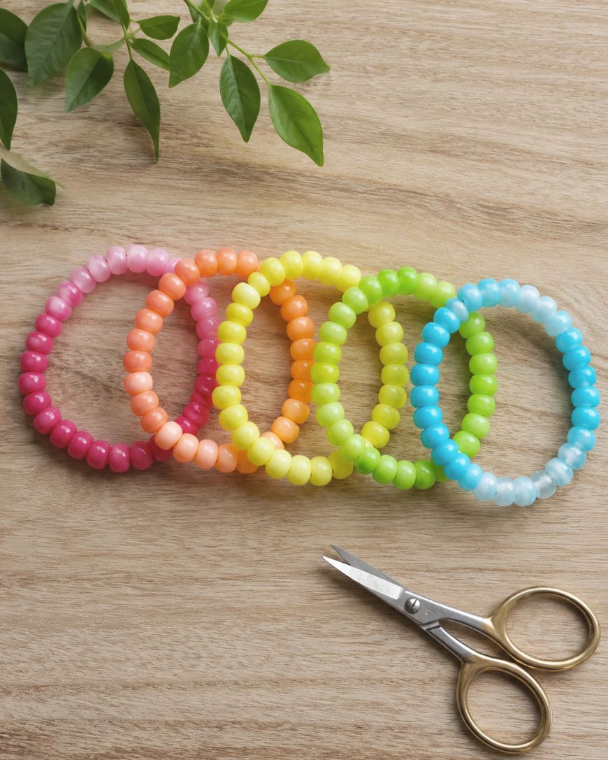 Six colorful bead bracelets are arranged in a row on a wooden surface, with a pair of small scissors below them and green leaves in the top left corner.