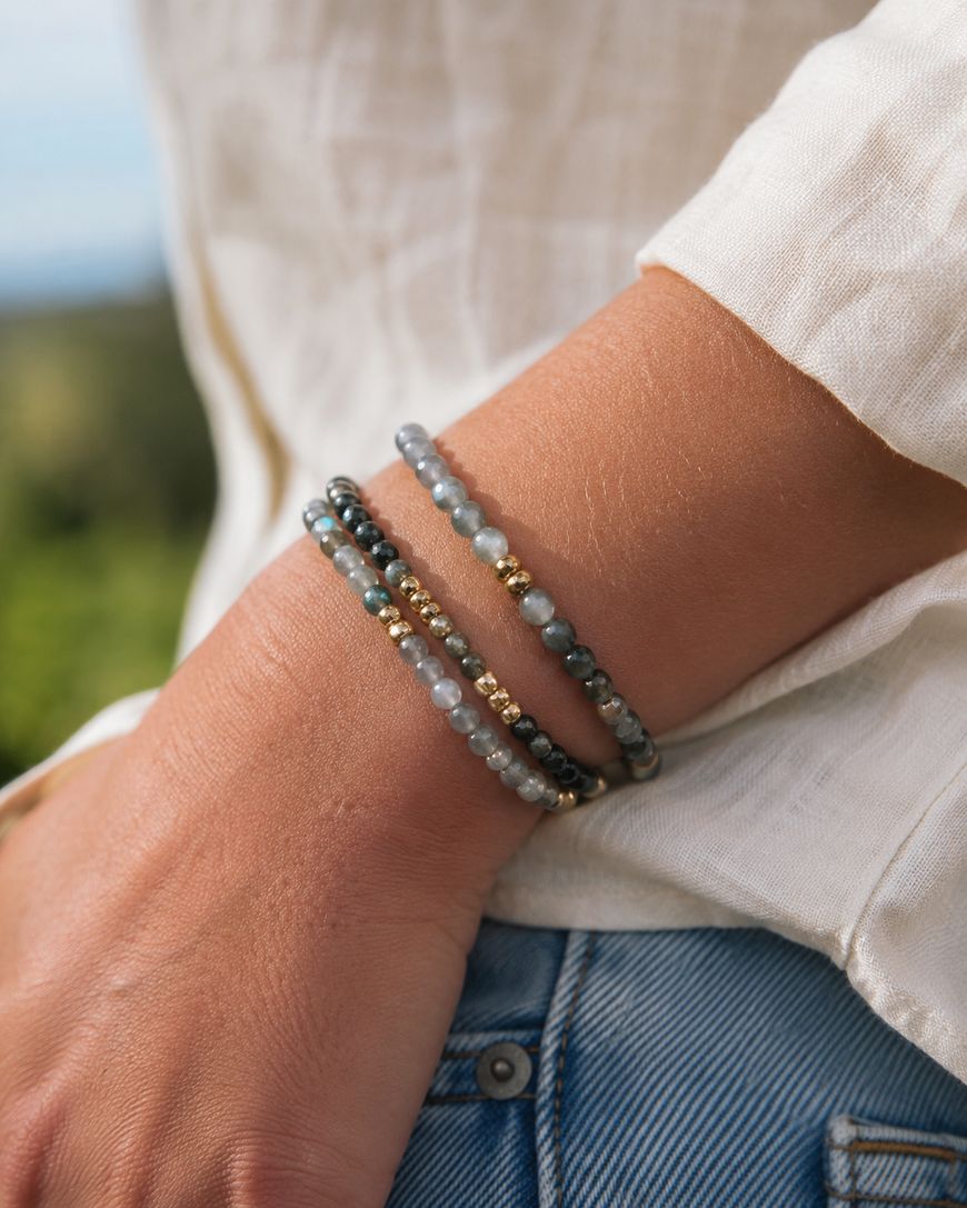 A person wearing a cream-colored long-sleeve shirt and blue jeans has three beaded bracelets with gold accents on their wrist, photographed outdoors.