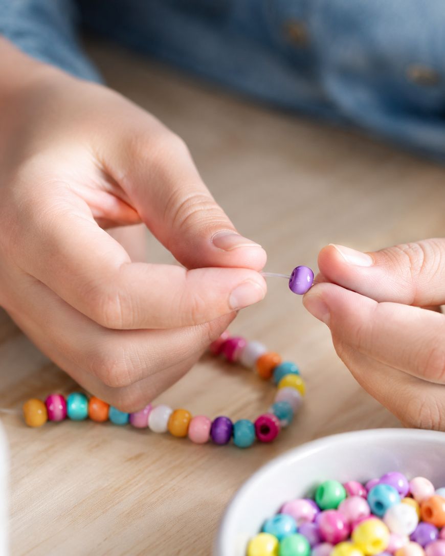 Close-up of hands stringing colorful round beads onto a cord. A finished part of the beaded necklace and a bowl filled with more multicolored beads are visible on a light wooden surface.
