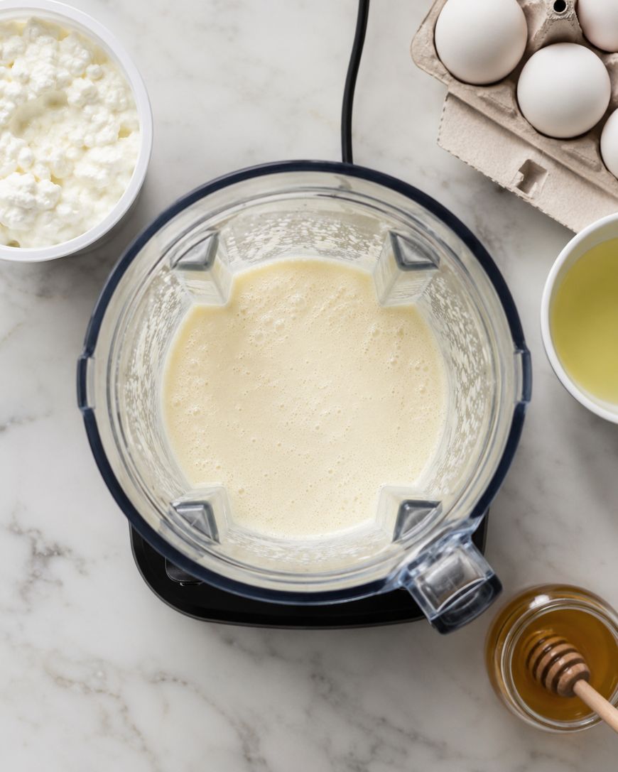 A blender with a creamy mixture inside sits on a marble counter, surrounded by a bowl of cottage cheese, eggs in a carton, a bowl of egg whites, and a jar of honey.