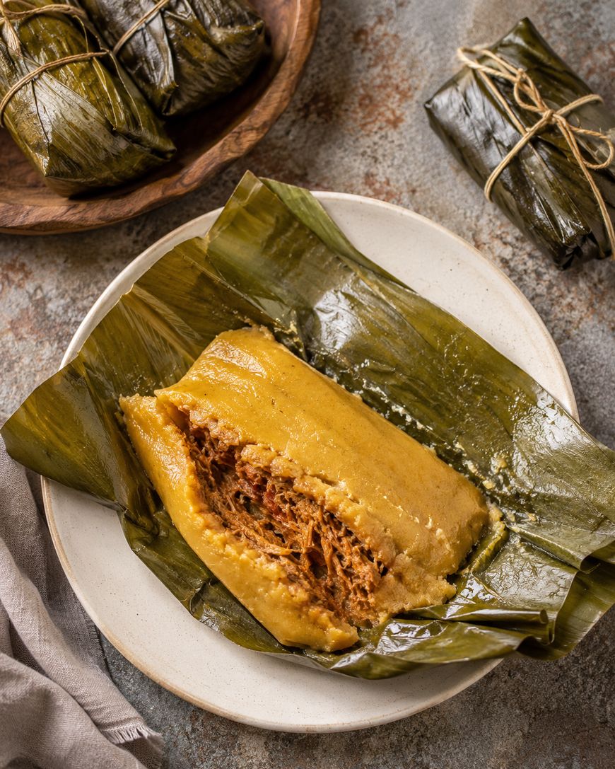A plate holds a tamal wrapped in a banana leaf, partially opened to reveal a savory meat filling. More wrapped tamales are seen in the background on a wooden plate and the table.