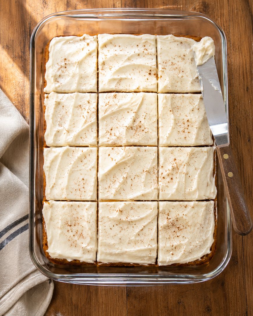 A glass baking dish with twelve frosted cake squares, lightly sprinkled with spice, sits on a wooden table. A metal spatula with a wooden handle rests on the edge of the dish.