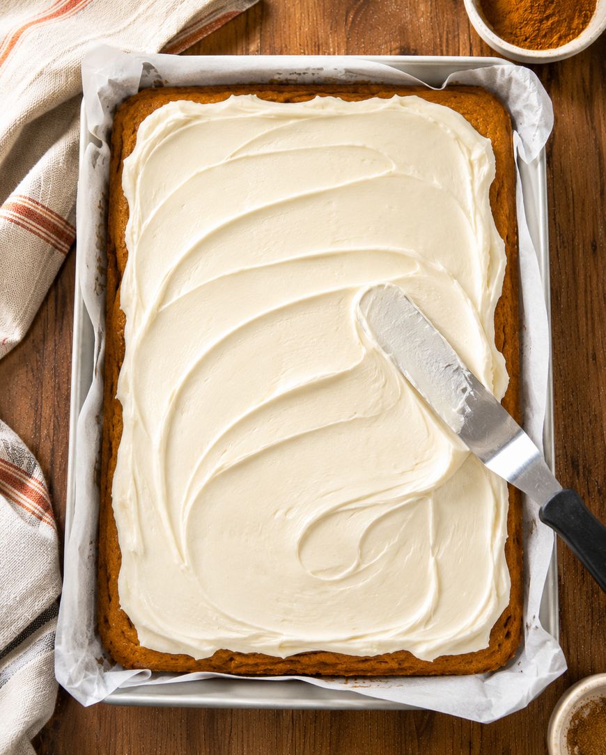 A rectangular cake with creamy white frosting is being spread with an offset spatula in a parchment-lined baking pan on a wooden table.