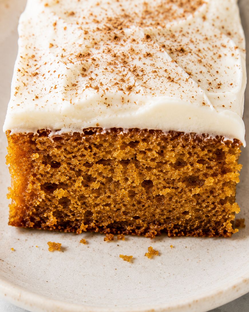 A close-up of a slice of moist pumpkin cake topped with creamy white frosting and a sprinkle of cinnamon, served on a white plate.