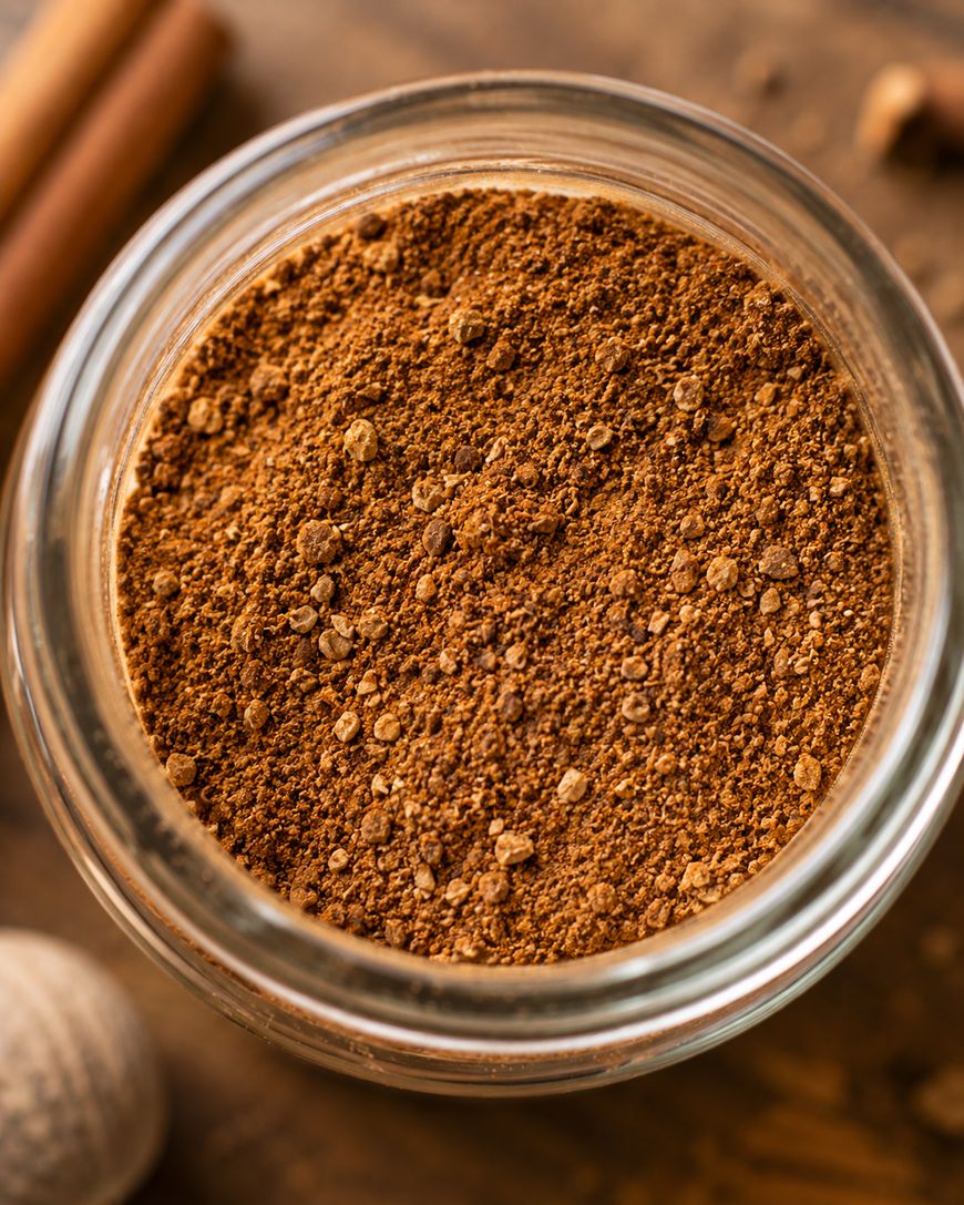 A close-up of a glass jar filled with ground brown spices, likely a blend such as pumpkin pie spice, viewed from above on a wooden surface.