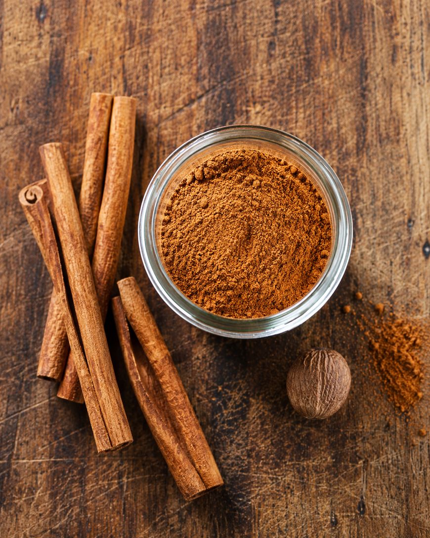 A glass bowl of ground cinnamon sits on a wooden surface next to several cinnamon sticks and a whole nutmeg.