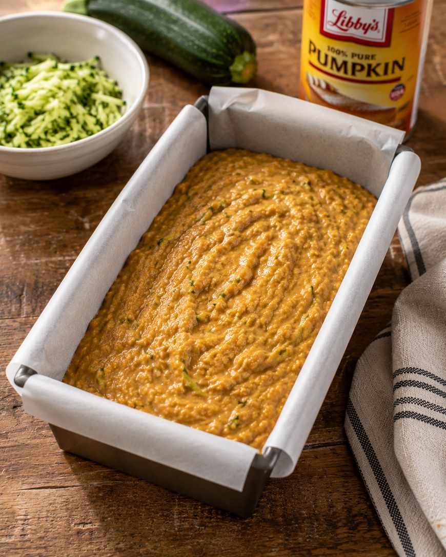 A loaf pan filled with pumpkin bread batter sits on a wooden table. Nearby are a can of pumpkin, a bowl of shredded zucchini, a whole zucchini, and a striped cloth napkin.