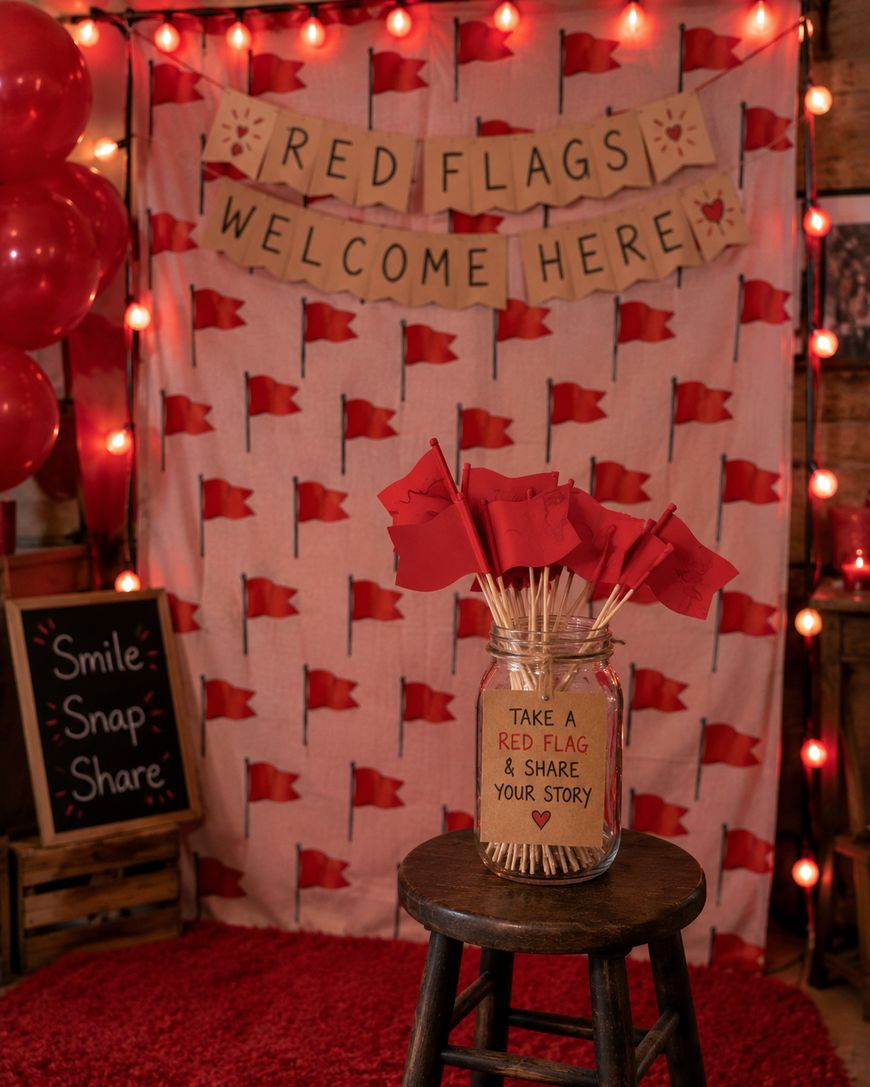 A photo booth with red flag decor, a Red Flags Welcome Here banner, red lights, and a jar of red flag props on a stool. A chalkboard says Smile-Snap-Share. The background features a red flag pattern.