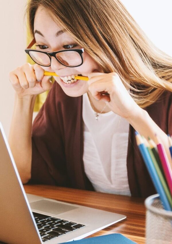 A woman wearing glasses sits at a desk, biting a yellow pencil and looking at a laptop screen with a stressed expression. Colored pencils are in a holder on the desk in the foreground.
