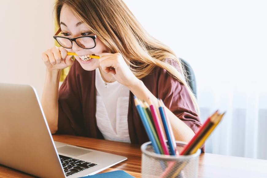 A woman wearing glasses sits at a desk with a laptop, biting a pencil and looking stressed. A container of colored pencils and notebooks are on the desk, and sunlight comes through a window in the background.