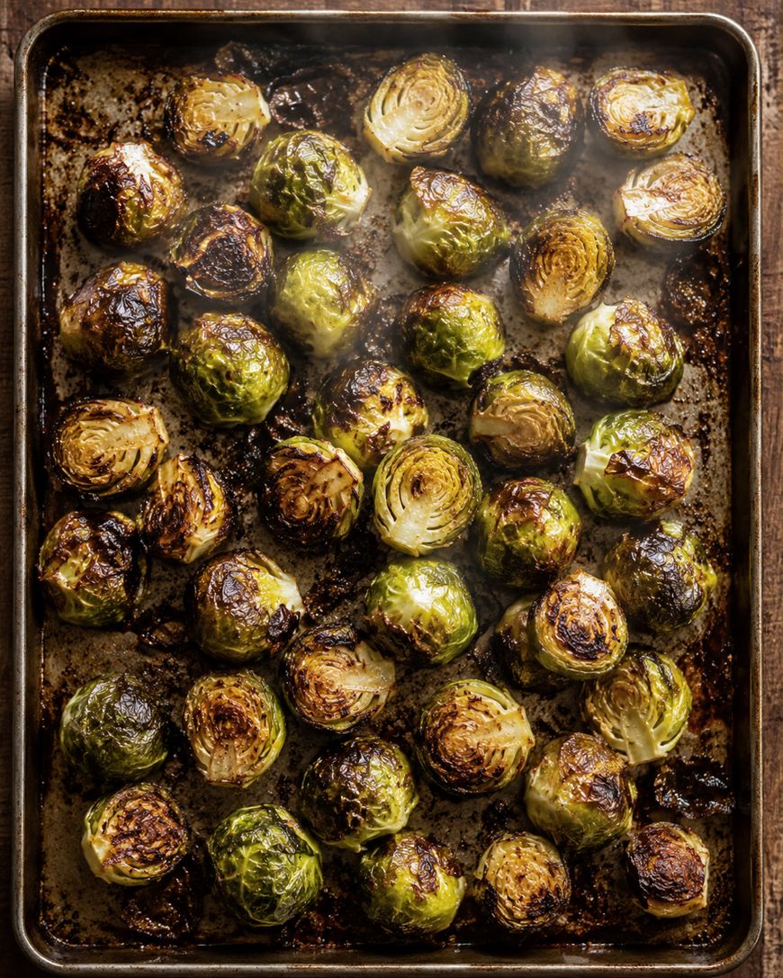A baking tray filled with roasted Brussels sprouts, browned and crispy on the edges, arranged in rows on a dark baking sheet. Steam rises from the hot vegetables.