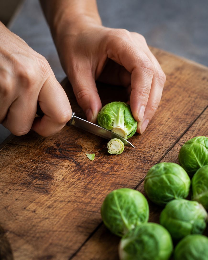 A person slicing the stem off a Brussels sprout with a small knife on a wooden cutting board, with several whole Brussels sprouts nearby.