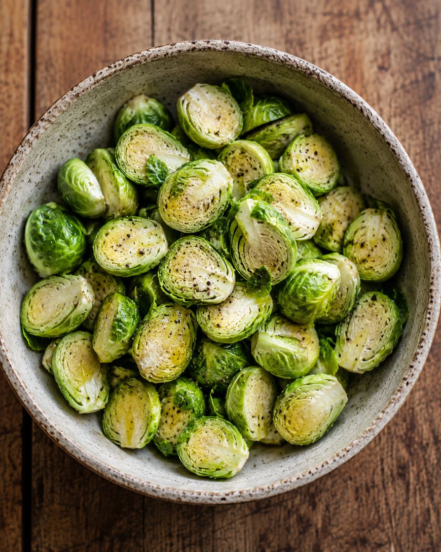 A bowl of halved Brussels sprouts seasoned with salt, pepper, and oil sits on a wooden surface.