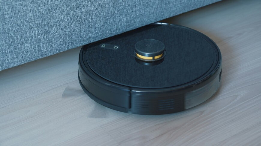 A black robotic vacuum cleaner operates on a light-colored wooden floor, cleaning underneath a gray fabric sofa.
