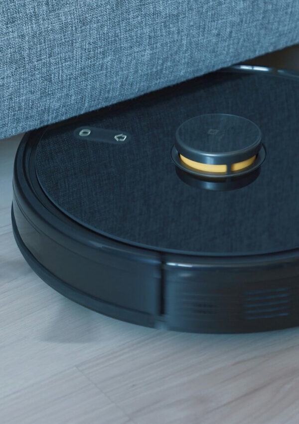 A black robotic vacuum cleaner is cleaning a light wood floor, moving partially under a gray fabric sofa.