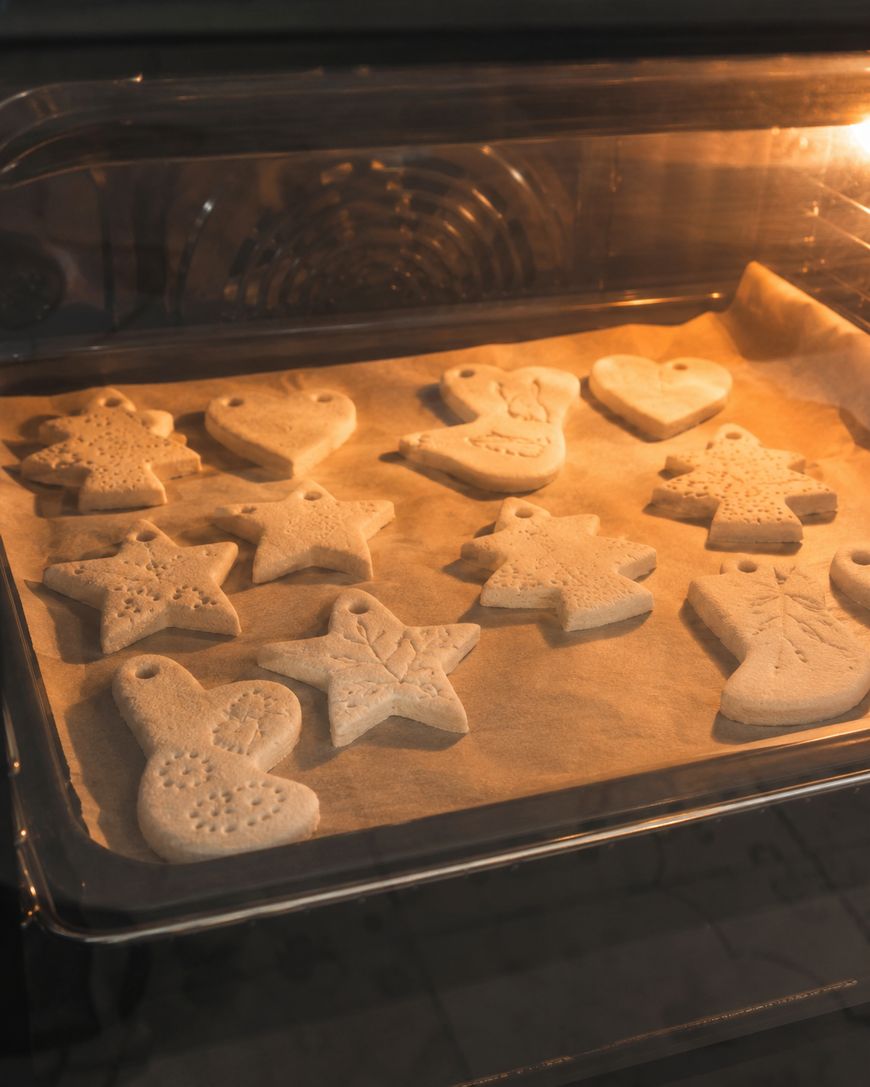 Unbaked, shaped salt dough ornaments, including stars, hearts, and birds, are on a baking tray lined with parchment paper inside an oven.