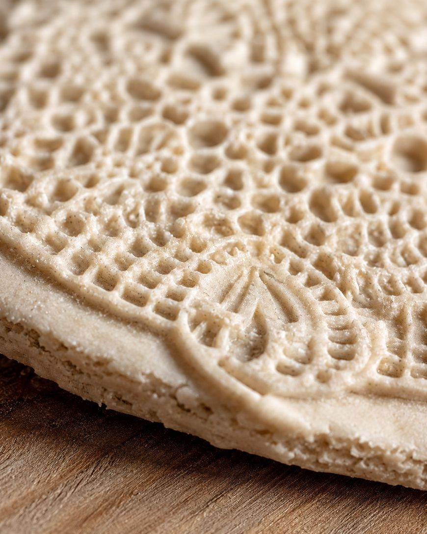 A close-up of a round, beige, intricately patterned cookie or wafer resting on a wooden surface. The texture and detailed design are clearly visible.