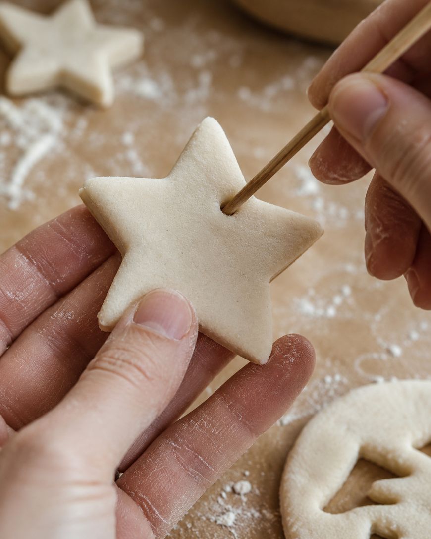A person uses a wooden skewer to poke a hole in a raw, star-shaped cookie dough piece. Flour is visible on the hands and the surface below.